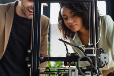 young interracial colleagues looking at 3D printer producing plastic figure in modern office