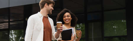 smiling young interracial couple with paper cups looking at each other and walking near building,...