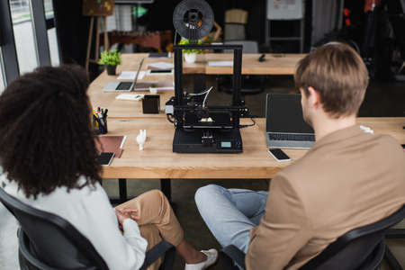 back view of young interracial designers sitting near 3D printer in modern open spaceの写真素材