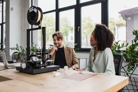 young interracial designers sitting near 3D printer, plastic figures and laptop in modern open space