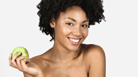 Smiling african american woman with clean skin looking at camera and holding green apple isolated on...