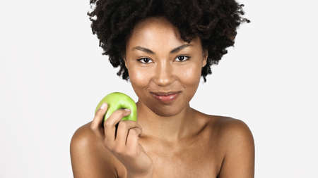 Smiling african american woman with clean skin holding whole apple isolated on gray