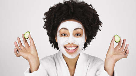 Smiling african american woman with mask on face holding slices of cucumber isolated on grayの写真素材