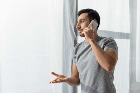 Side view of man pointing with hand while talking on smartphone near window at home