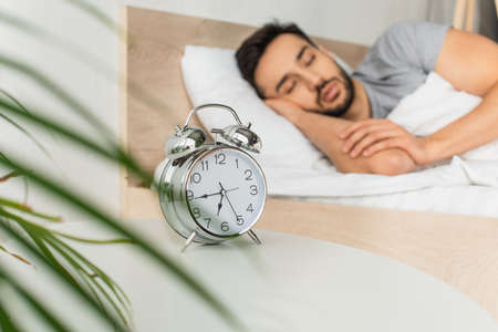 Alarm clock near blurred plant and man sleeping on bed
