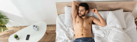 Top view of shirtless man lying near alarm clock and smartphone on bedside table, bannerの写真素材