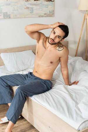 Shirtless man in pants looking at camera while sitting on bedの写真素材