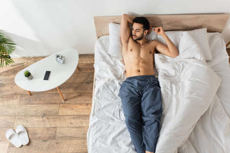 Top view of shirtless man lying on bed near smartphone with blank screen and alarm clock on bedside tableの写真素材