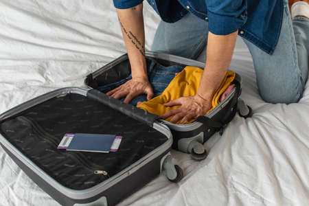 Cropped view of tourist pressing clothes in suitcase near passport with boarding pass on bed