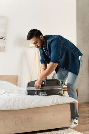 Bearded traveler fastening suitcase on bed at homeの写真素材