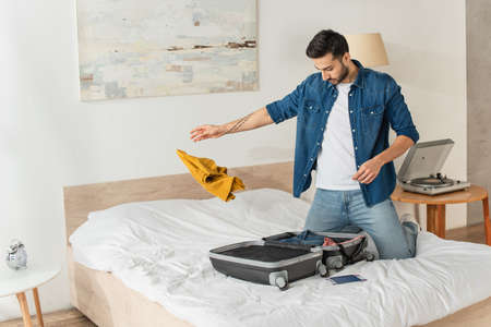 Man throwing clothes near suitcase and passport on bedの写真素材