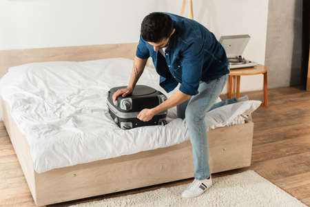 Young man zipping suitcase on bed at homeの写真素材