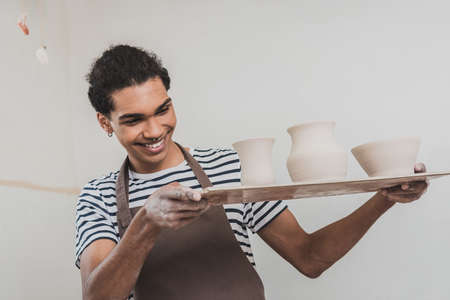 smiling young african american man looking at clay pots on tray in potteryの写真素材