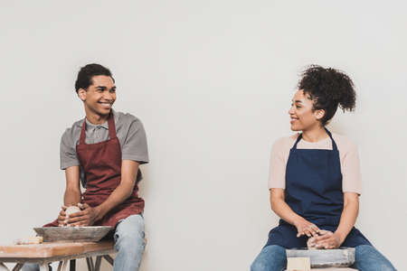 smiling young african american couple in aprons modeling wet clay on wheels and looking at each other in potteryの写真素材