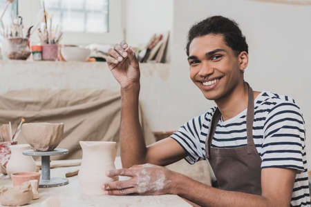 smiling young african american man looking at camera near clay pot and stick in hand in potteryの写真素材