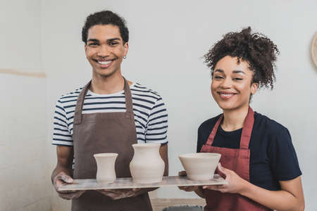 smiling young african american couple holding clay pots on tray and looking at camera in potteryの写真素材