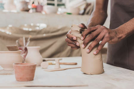 partial view of young african american man working with piece of clay on table with equipment in...