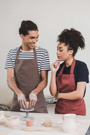 smiling young african american man making clay pot near woman with hand near face in potteryの写真素材