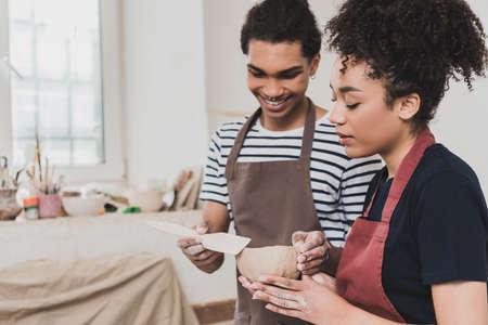 smiling young african american man standing with clay pot and spatula near focused woman in potteryの写真素材