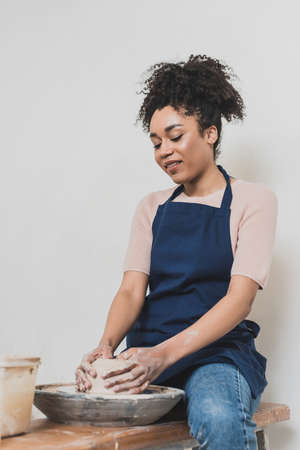 cheerful young african american woman sitting on bench and modeling wet clay on wheel with hands in...