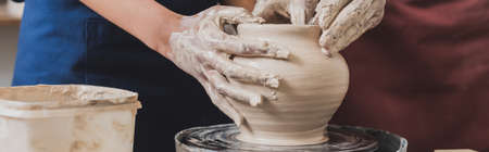 partial view of young african american couple shaping wet clay pot on wheel with hands in pottery, bannerの写真素材