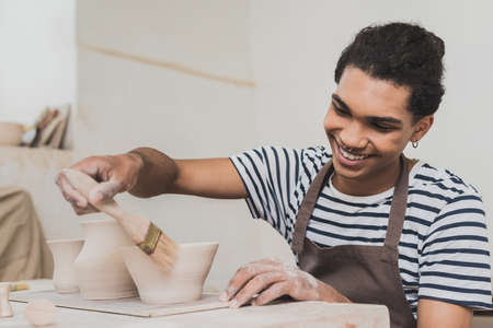 smiling young african american man glazing clay pots with brush in potteryの写真素材