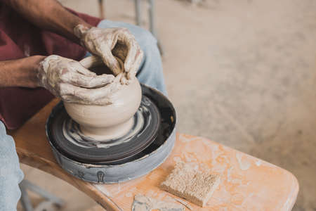 partial view of male african american hands shaping wet clay pot on wheel near sponge and scraper in...