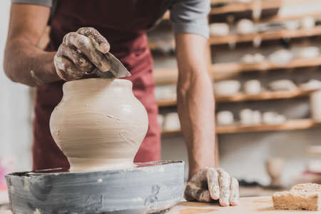 partial view of young african american man in apron shaping wet clay pot on wheel with scraper in...