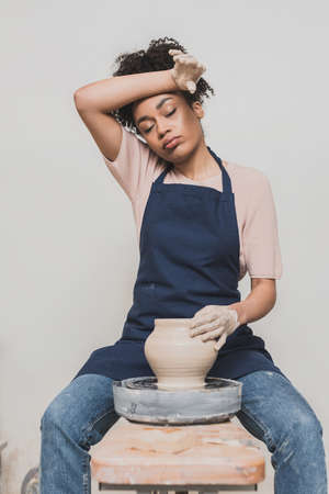 tired young african american woman in apron sitting on bench and shaping wet clay pot with hand near head in potteryの写真素材