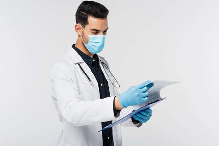 Arabian doctor in latex gloves and medical mask holding clipboard isolated on grayの写真素材