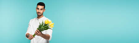 Young arabian man holding tulips isolated on blue, bannerの写真素材