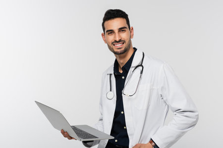 Cheerful arabain doctor holding laptop and looking at camera isolated on grayの写真素材