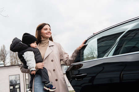 pleased woman looking away while holding little son near car outdoorsの写真素材