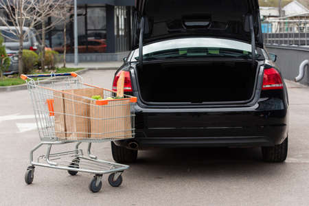 shopping cart with fresh grocery near black car with open trunkの写真素材