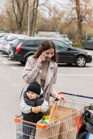young woman talking on smartphone near purchases and toddler son in shopping cartの写真素材