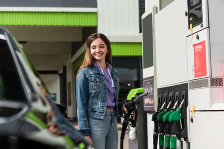 smiling woman in denim clothes holding gasoline pistol near blurred carの写真素材