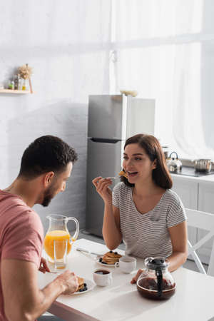 happy young woman eating pieces of pancakes on fork with boyfriend in kitchenの写真素材