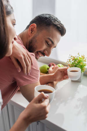 smiling young woman holding white cup with coffee and touching boyfriend near window in kitchenの写真素材