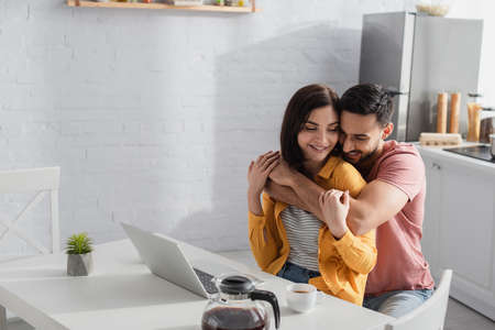 smiling young man hugging girlfriend with laptop in kitchenの写真素材