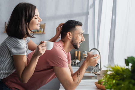 positive young woman holding white cup with coffee and touching boyfriend near window in kitchen