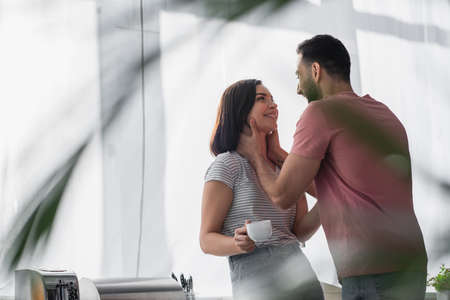 smiling young man touching neck of girlfriend with white coffee cup in modern kitchen