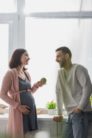positive young man standing near pregnant woman with apple in kitchenの写真素材