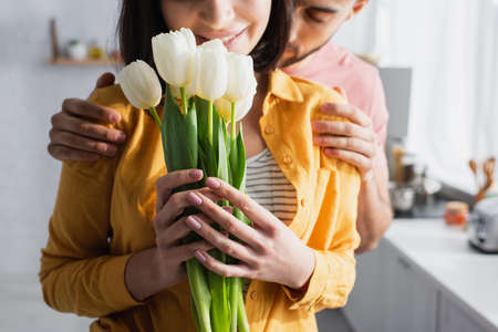 partial view of young man hugging girlfriend with bouquet of flowers in kitchenの写真素材