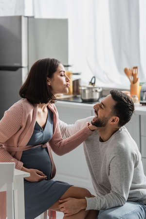 smiling young man sitting near pregnant woman with pouting lips in kitchenの写真素材