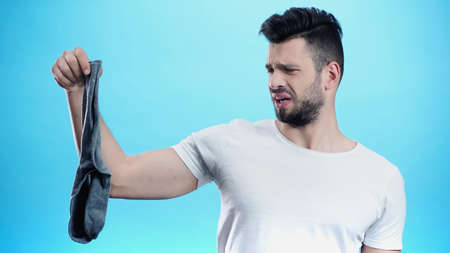 disgusted man holding stinky sock isolated on blue