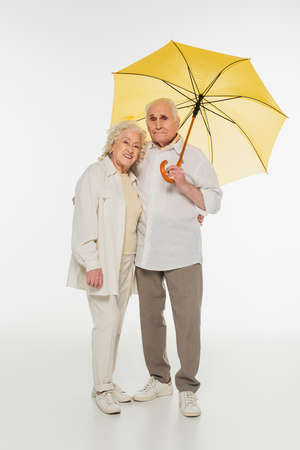 smiling elderly couple in casual clothes standing with yellow umbrella on whiteの写真素材