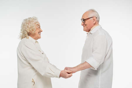 smiling elderly couple in casual clothes holding hands and looking at each other isolated on whiteの写真素材