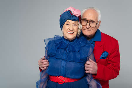 smiling elderly man in red blazer hugging wife in blue dress and turban isolated on grayの写真素材