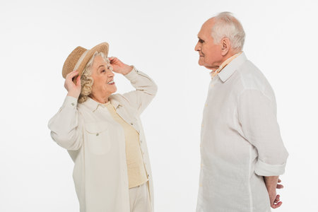 positive elderly woman adjusting hat on head and looking at husband isolated on whiteの写真素材