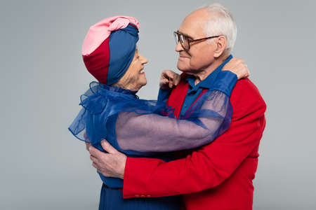 smiling elderly couple in blue dress and red blazer hugging isolated on grayの写真素材